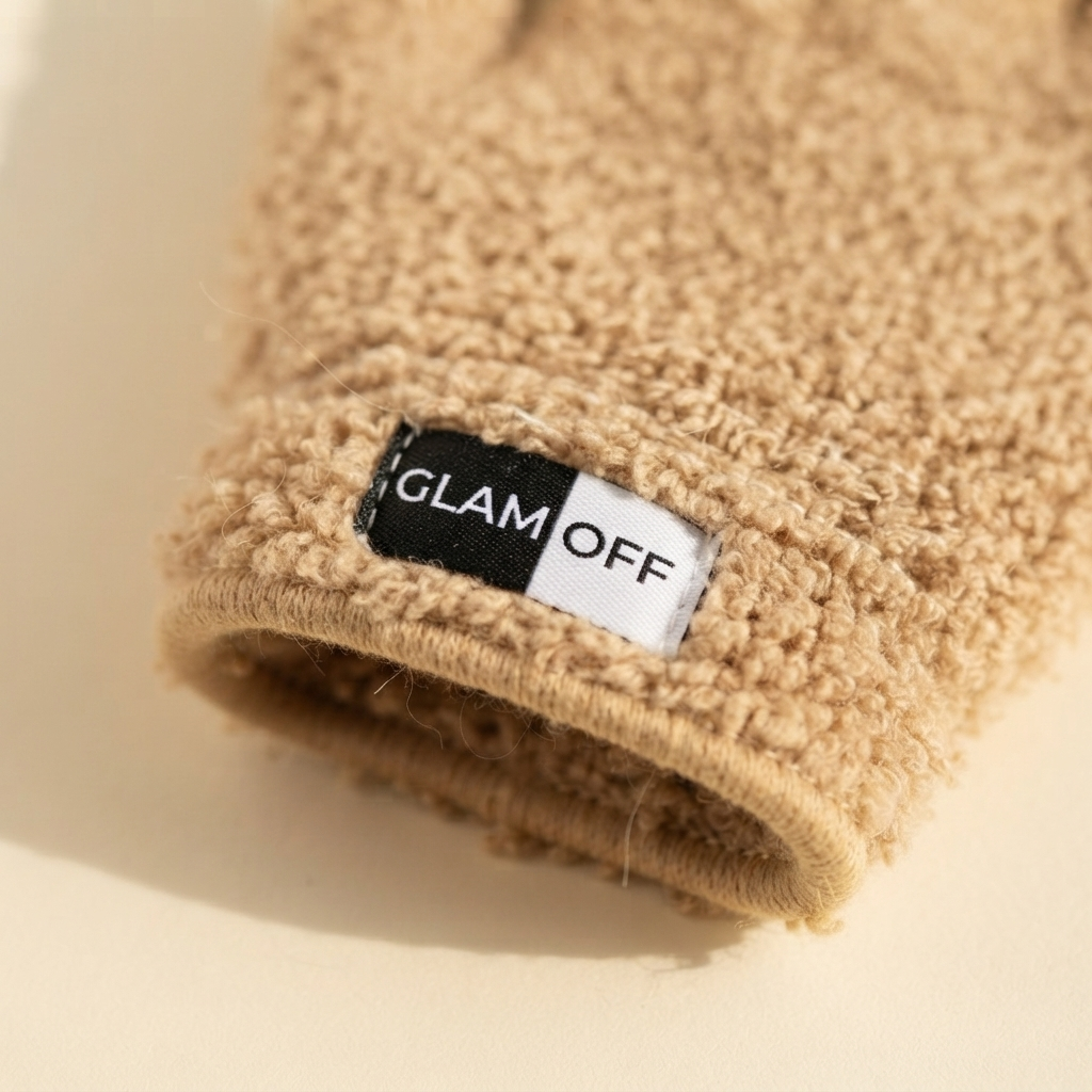 Close-up of a beige textured microfibre glove with a black and white brand label reading 'GLAM OFF', set against a warm cream background.