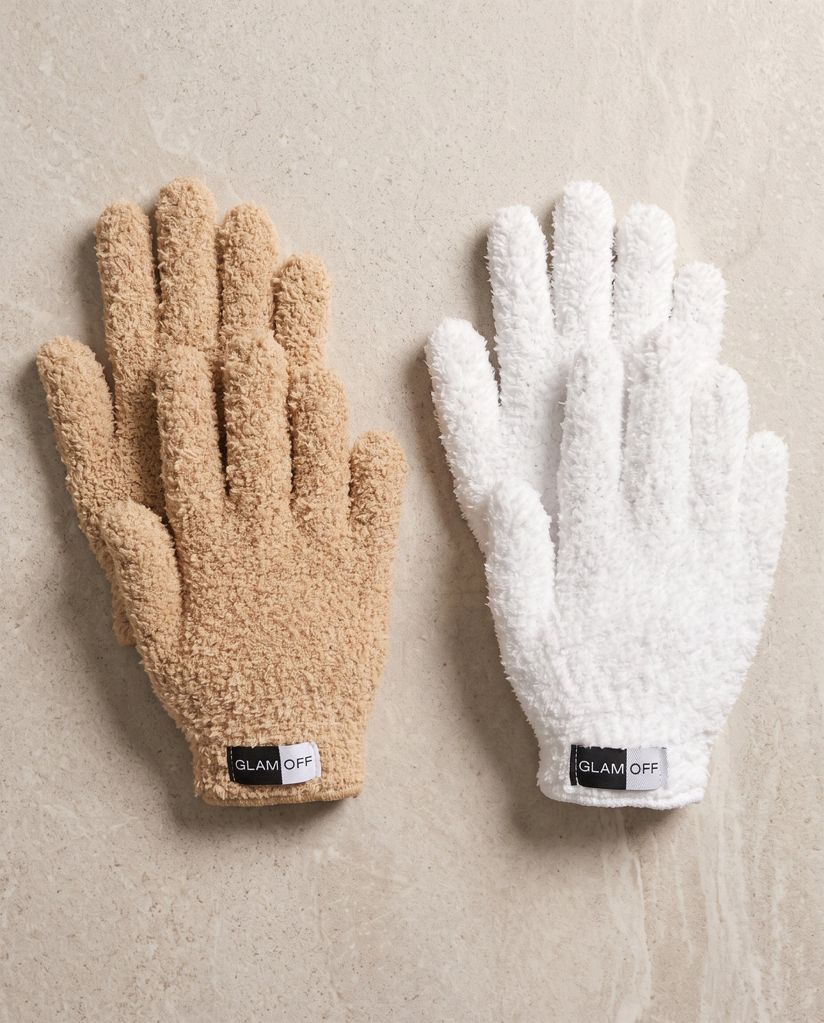 Two plush microfibre gloves laid flat on a light beige stone surface, one in warm beige (oat) and one in white, each with a small black GLAM OFF label at the cuff.