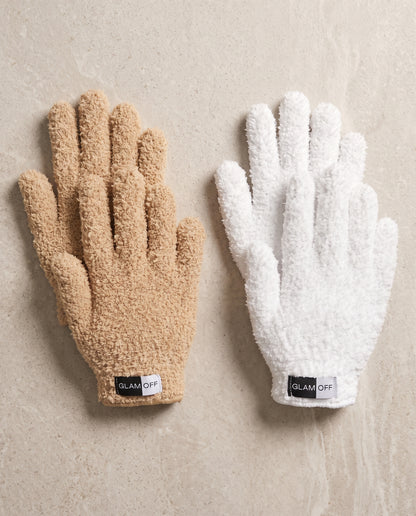 Two plush microfibre gloves laid flat on a light beige stone surface, one in warm beige (oat) and one in white, each with a small black GLAM OFF label at the cuff.