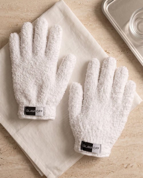 A pair of white textured plush microfibre gloves with 'GLAM OFF' labels, laid flat on a beige towel over a light stone surface.