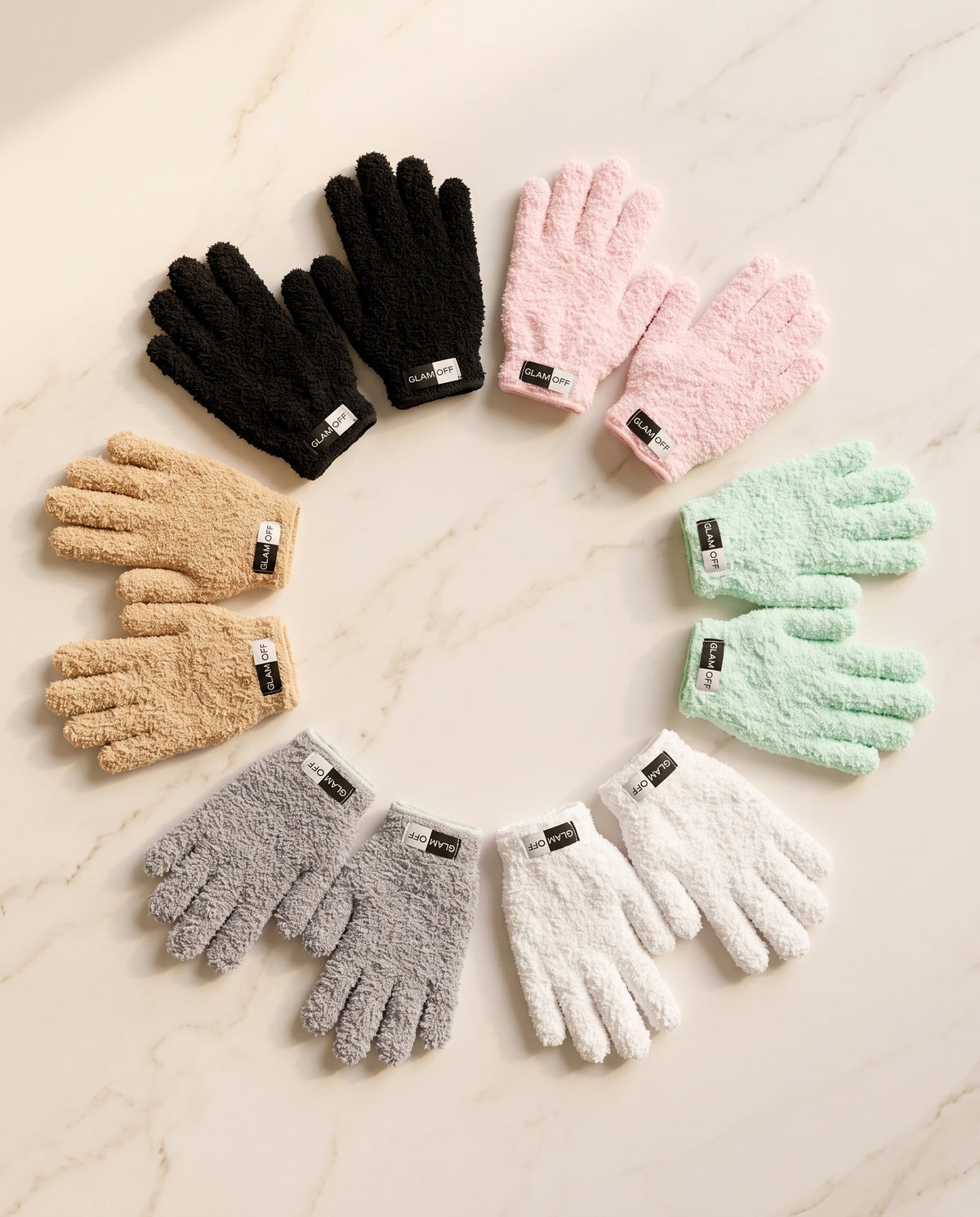 Six pairs of fuzzy microfibre gloves arranged in a circular pattern on a light beige marble background. The gloves come in black, light pink, tan (oat), mint green, light gray/grey, and white.