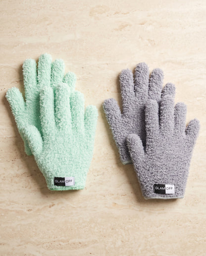 Two pairs of plush microfibre gloves, one mint green and one heather gray, displayed flat on a light beige stone surface. Each pair has a black and white 'GLAM OFF' brand tag at the cuff.
