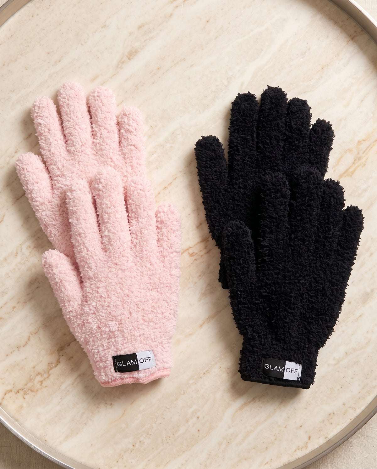 Two pairs of plush microfibre gloves, one light pink and one black, displayed side-by-side on a light beige marble tray. Both gloves feature a textured fuzzy fabric and a black cuff label with the brand name 'GLAM OFF'.