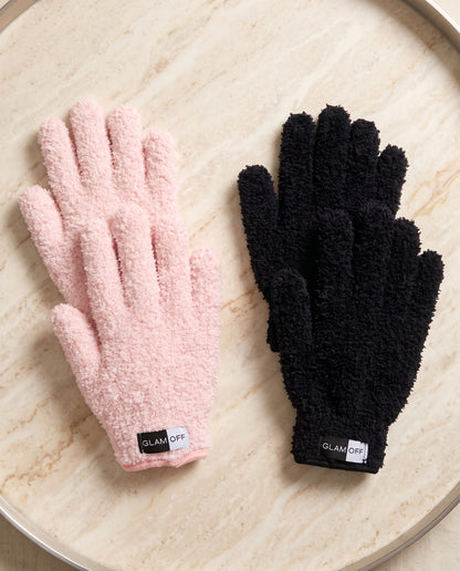 Two pairs of plush microfibre gloves, one light pink and one black, displayed side-by-side on a light beige marble tray. Both gloves feature a textured fuzzy fabric and a black cuff label with the brand name 'GLAM OFF'.